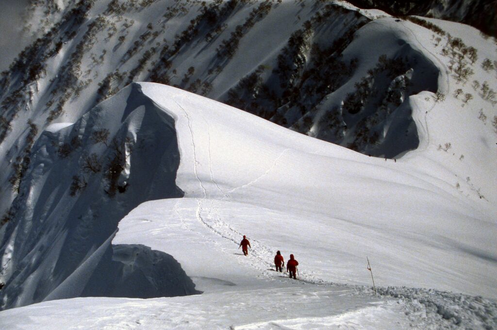 Three people walking in a line along a narrow snowy path on a steep white mountain, surrounded by deep snow and rugged terrain under a pale sky.