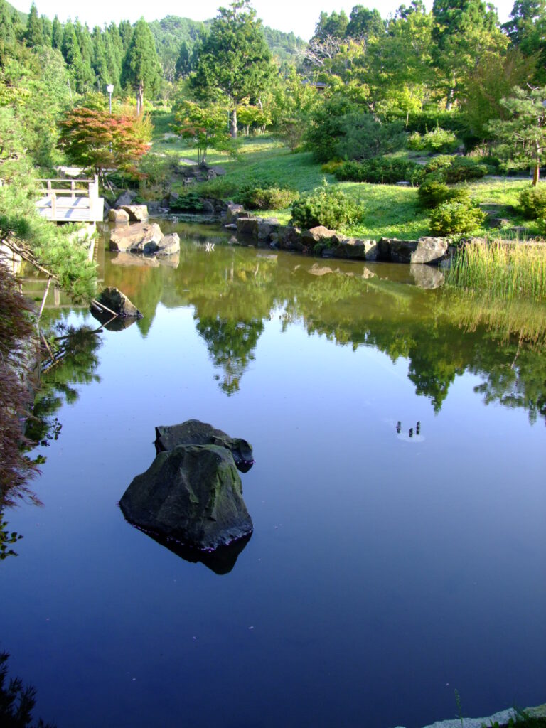 A quiet pond in a nature park in Hakodate, Hokkaido. A small wooden water mill stands near the edge of the water. Green mountains and trees are reflected clearly on the still surface of the pond, along with a dark blue sky. A single rock rests quietly in the water, emphasizing the calm and stillness of the scene.