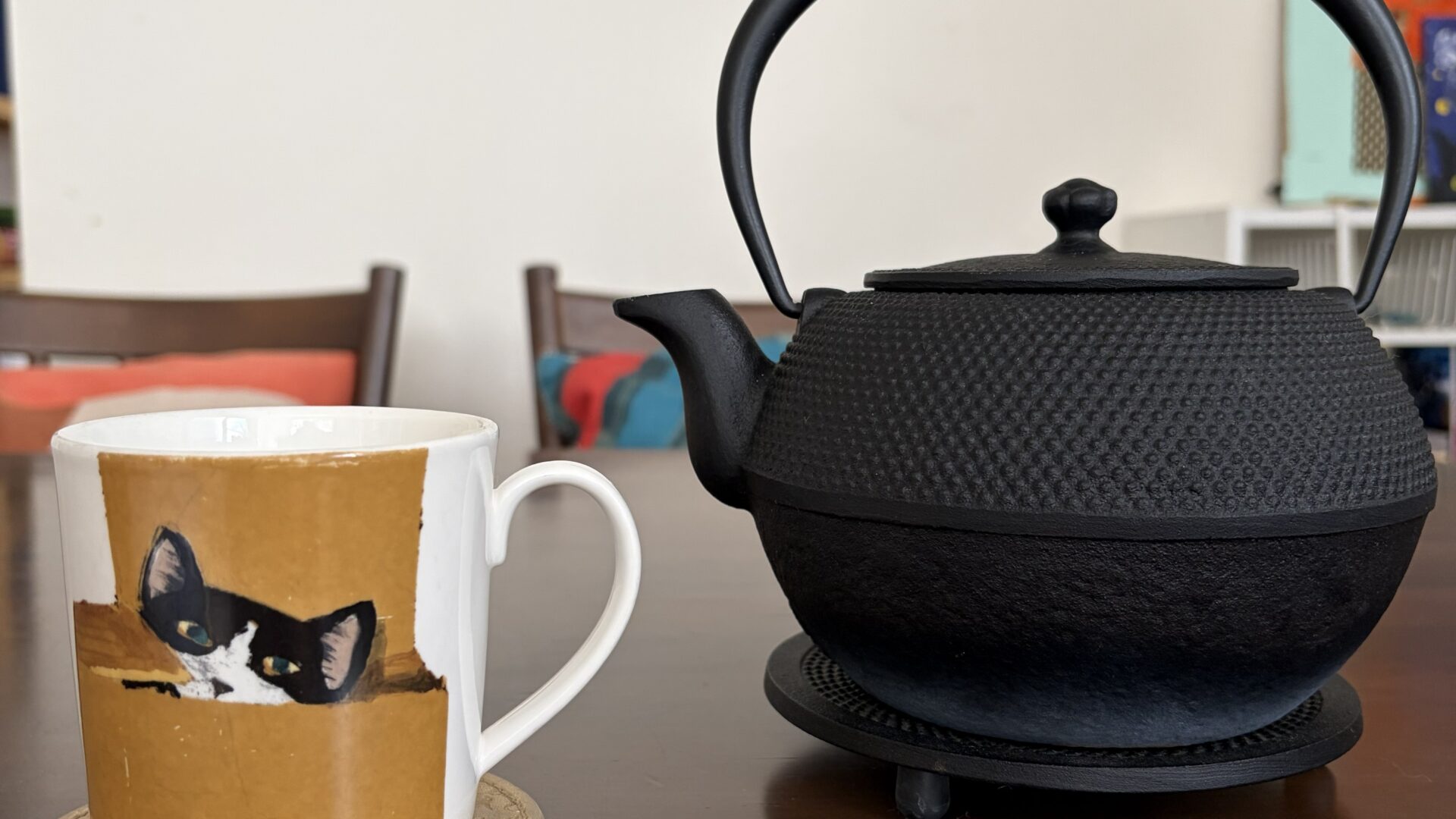 A simple tea setup on a clean dining table: a cup beside a black Nambu iron kettle (tetsubin), with small decorative items arranged in the background.
