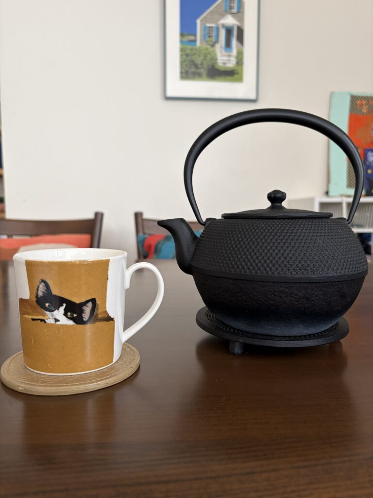 A simple tea setup on a clean dining table: a cup beside a black Nambu iron kettle (tetsubin), with small decorative items arranged in the background.